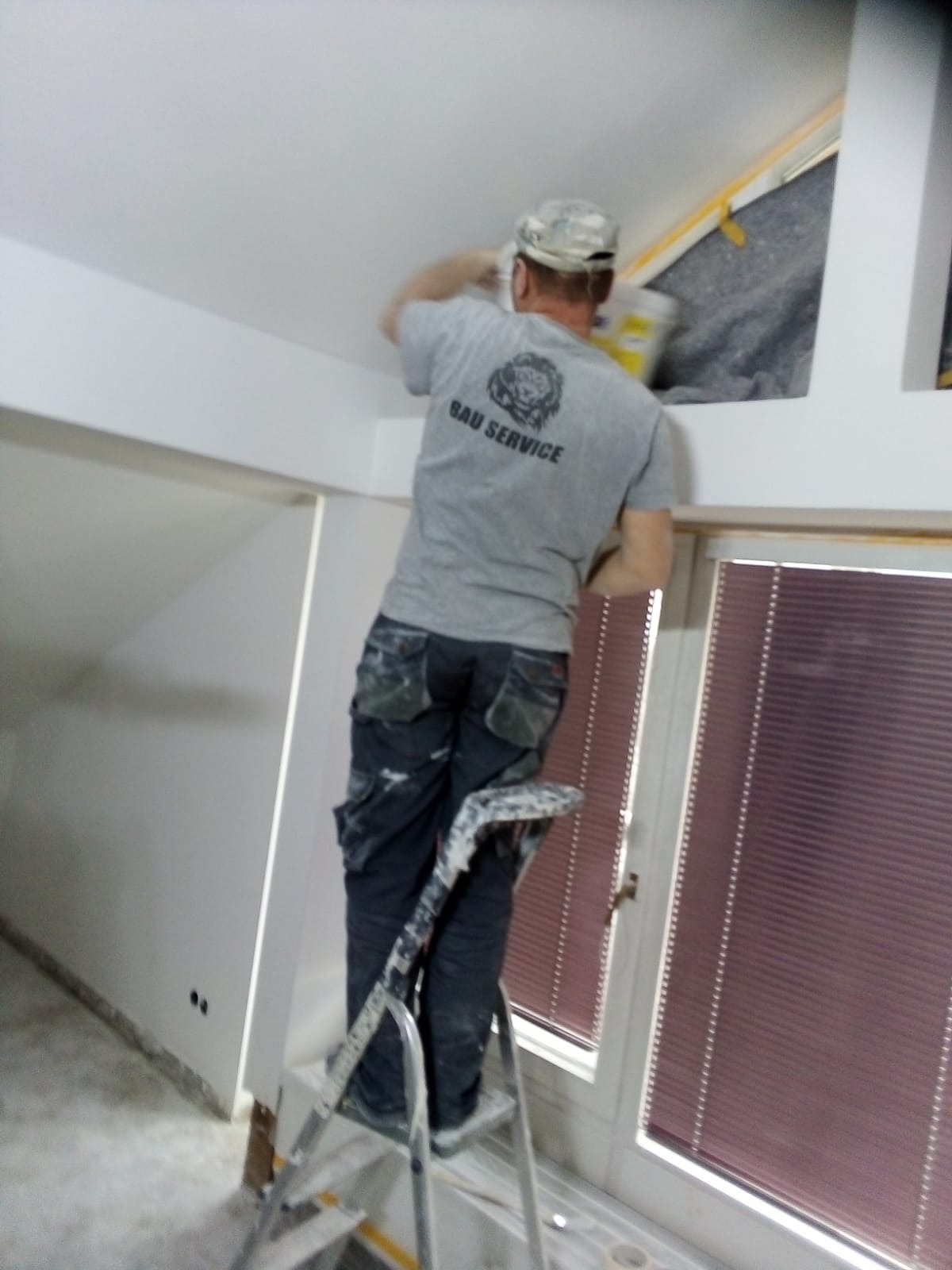 Mężczyzna w koszulce z logo 'BAU SERVICE' maluje sufit stojąc na aluminiowej drabinie w pomieszczeniu z oknem z żaluzjami.