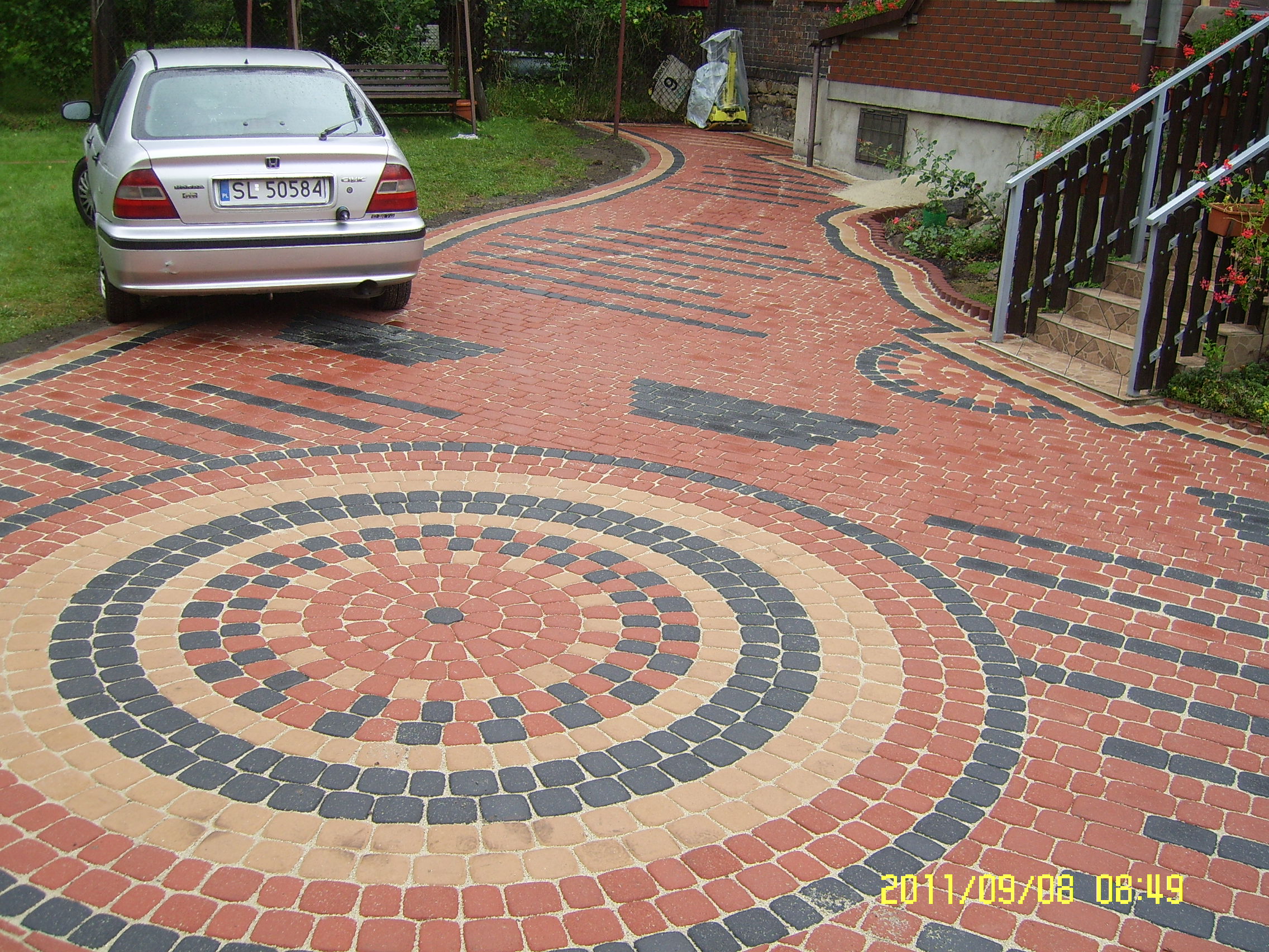 Ułożona kostka brukowa tworząca wzór koła i pasów, z zaparkowanym srebrnym samochodem osobowym w tle, obok budynku z cegły i schodów.
