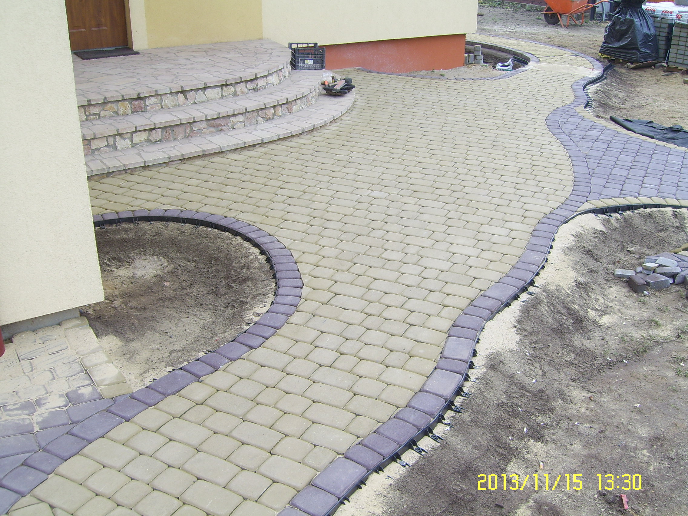 Kaskadowe schody wejściowe i wijąca się ścieżka z kostki brukowej w dwóch odcieniach szarości i beżu, obrzeża z tworzywa sztucznego stabilizujące krawędzie, widoczne narzędzia i materiały budowlane.