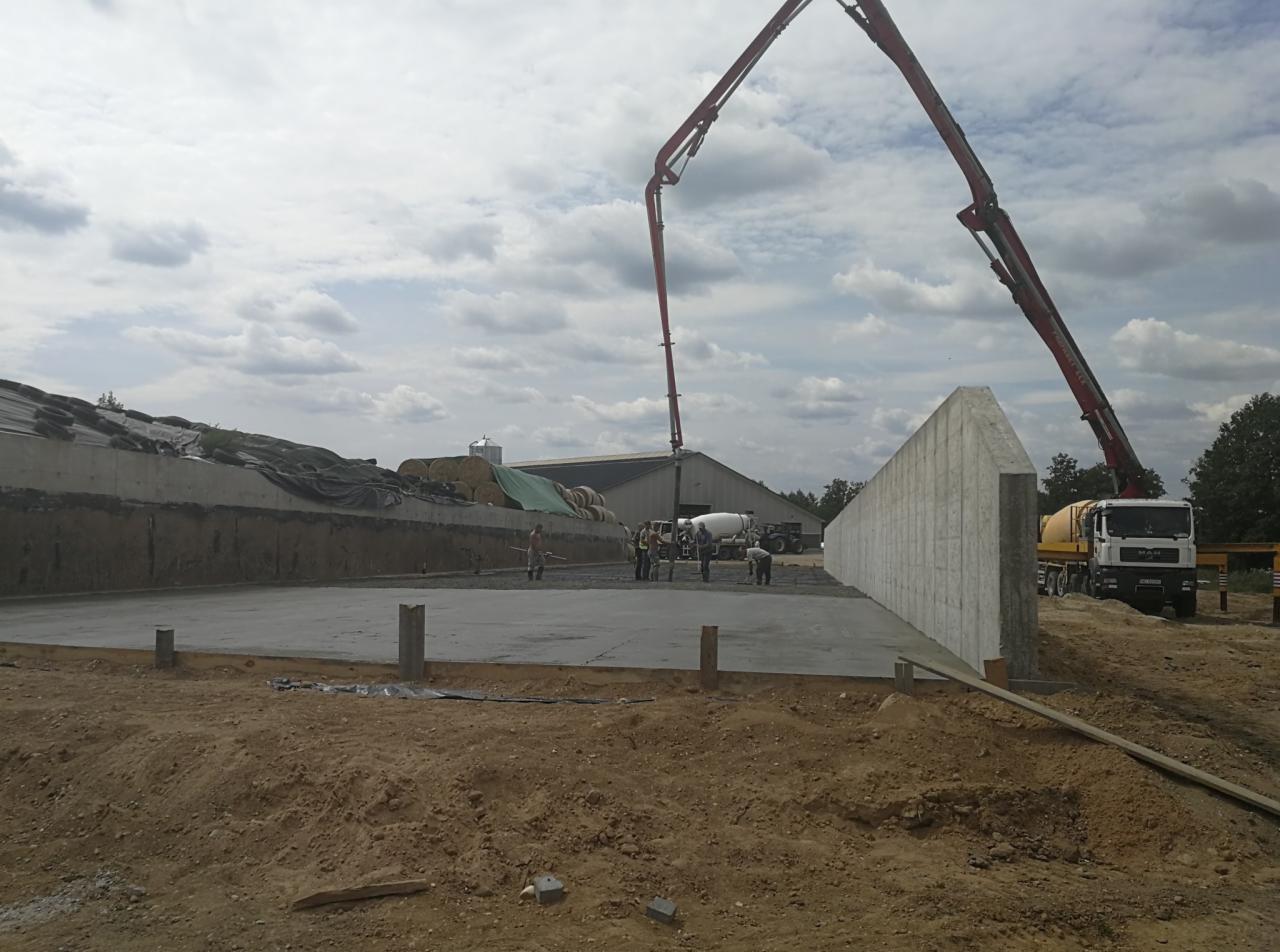 Wylewanie betonu na plac budowy z użyciem pompy do betonu, w tle budynek gospodarczy i betoniarka.