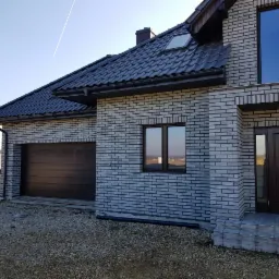 Nowy dom jednorodzinny z szarej cegły klinkierowej, z brązową bramą garażową i stolarką okienną, widok z zewnątrz na tle błękitnego nieba.