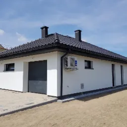 Widok z zewnątrz na nowo wybudowany, otynkowany na biało dom z szarą, automatyczną bramą garażową i klimatyzatorem na elewacji, z ciemną dachówką i czarnymi kominami.