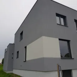 Nowoczesny dom z elewacją w odcieniach szarości, z fragmentem w kolorze złamanej bieli, widoczne okna z ciemnymi ramami, na tle pochmurnego nieba.