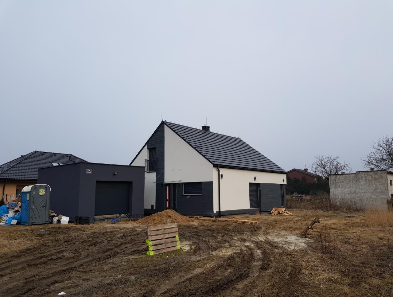 Nowoczesny dom jednorodzinny z grafitową bryłą garażu i białą elewacją, widoczny w trakcie prac wykończeniowych, z paletami i stertą piasku na błotnistym terenie.