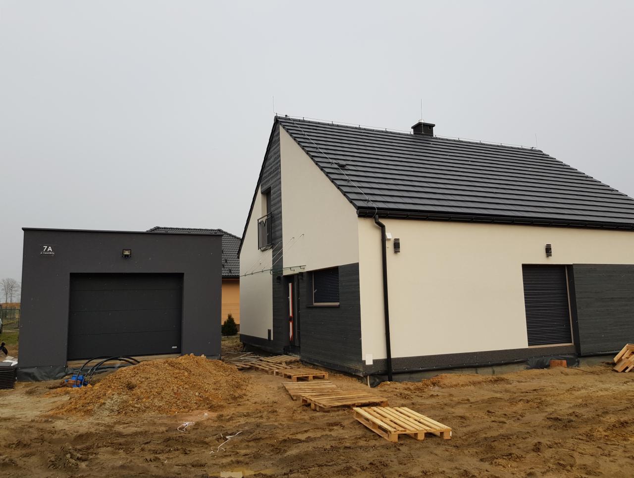 Nowoczesny dom jednorodzinny z garażem w trakcie wykańczania elewacji, widoczne palety na ziemi i sterta piasku, pochmurne niebo.