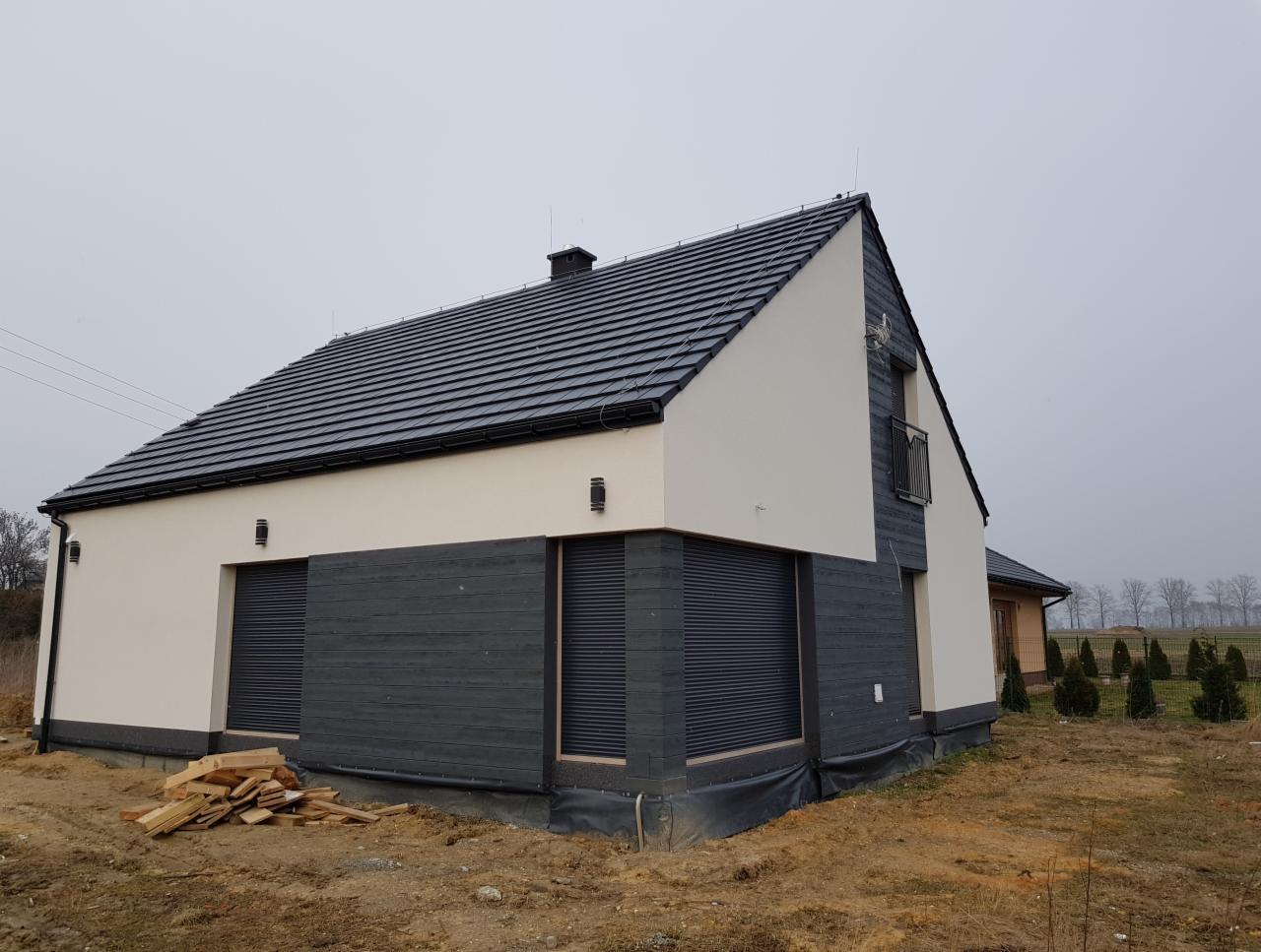 Nowoczesny dom jednorodzinny z elewacją w kolorach bieli i grafitu, z widoczną drewnianą okładziną i roletami zewnętrznymi, na działce w trakcie zagospodarowywania.