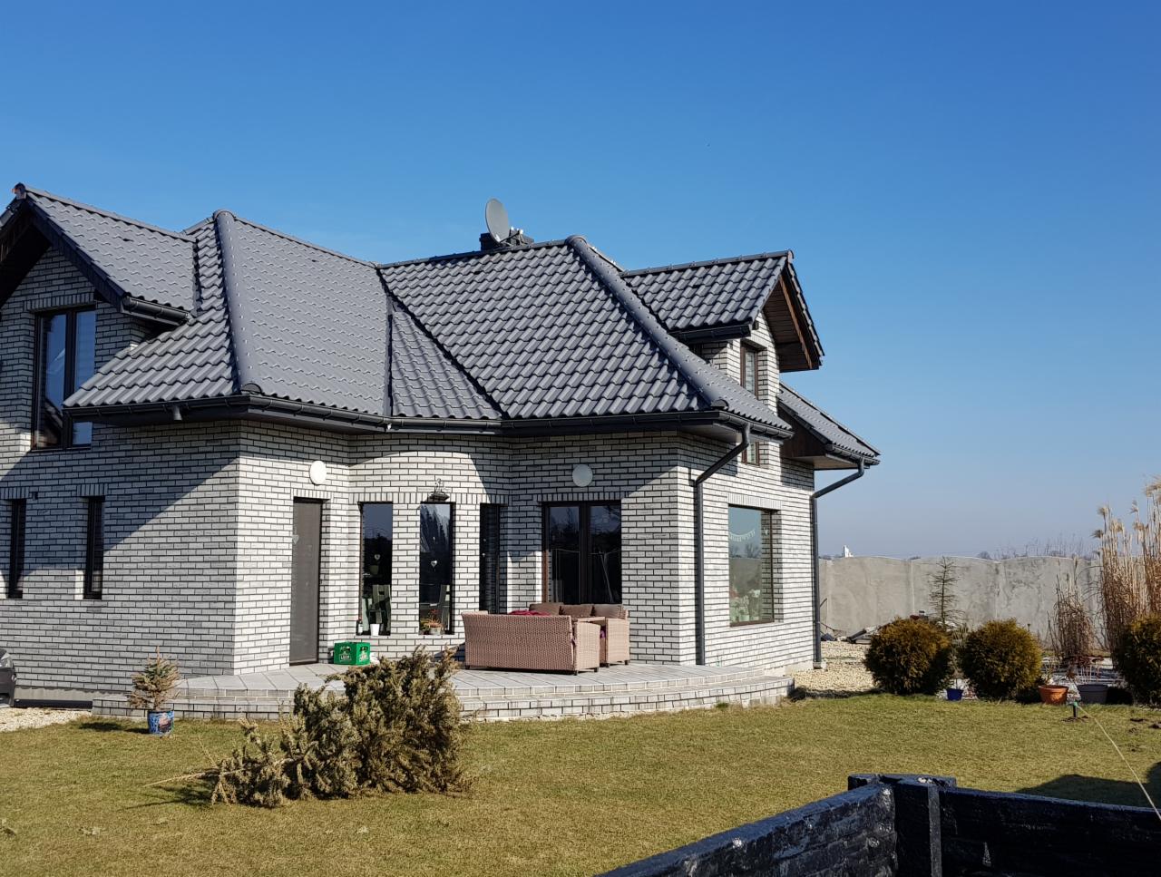 Dom jednorodzinny z jasnoszarej cegły z ciemnoszarą dachówką, widok z poziomu trawnika, w tle betonowe ogrodzenie i błękitne niebo.