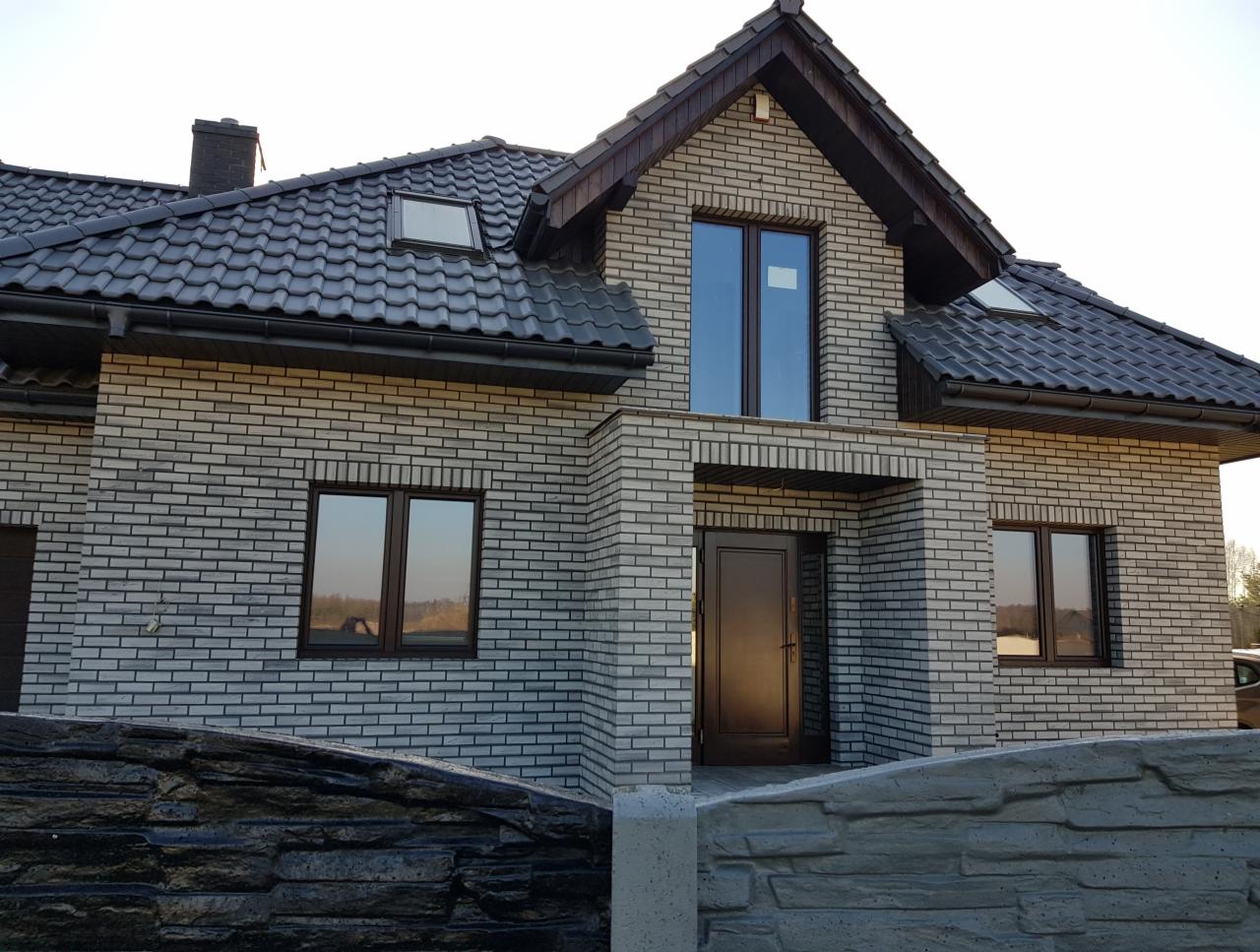 Nowoczesny dom jednorodzinny w Mikołowie z elewacją z szarej cegły klinkierowej, ciemną dachówką i brązowymi oknami, widok z poziomu ogrodzenia.