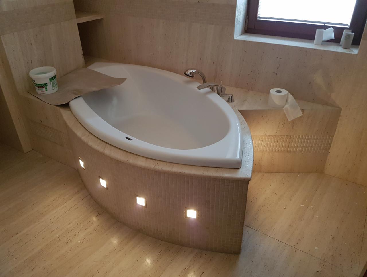 Nowoczesna wanna w obudowie z trawertynu z wbudowanymi punktami świetlnymi, widoczna rolka papieru toaletowego i wiaderko z masą uszczelniającą.