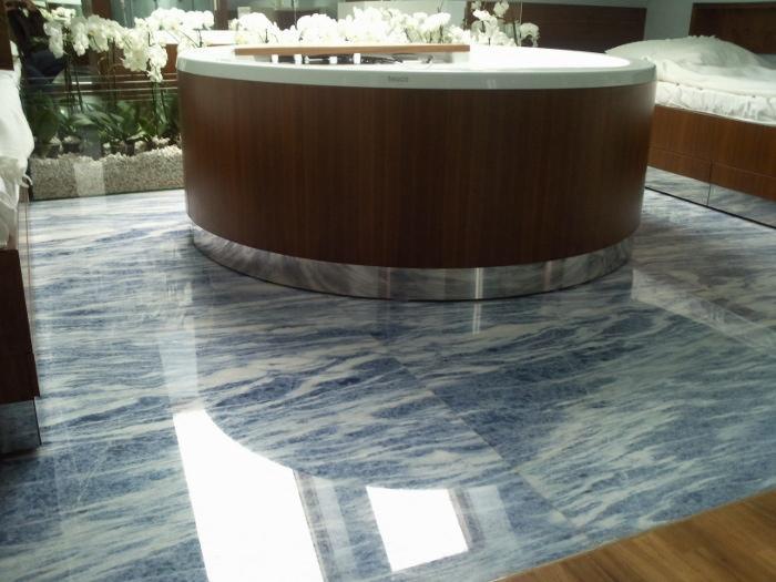 Szlifowana podłoga z niebieskiego kamienia z białymi żyłkami, odbijająca światło, z zaokrągloną obudową wanny z drewnopodobnym wykończeniem i metalicznym paskiem u podstawy, w tle rośliny i łóżko.