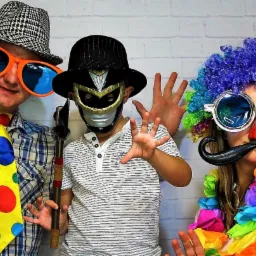 Trzy osoby w przebraniach pozują na tle białej ściany z cegieł: mężczyzna w kraciastej koszuli i dużych okularach, dziecko w masce superbohatera i kobieta w kolorowej peruce i okularach z wąsami.