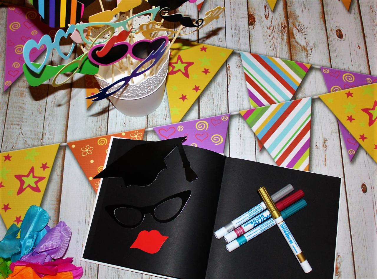 Przygotowania do sesji zdjęciowej: otwarty czarny album z wyciętymi elementami (kapelusz absolwenta, okulary, usta), kolorowe markery, rekwizyty (okulary, wąsy) w ozdobnym wiaderku i girlanda...