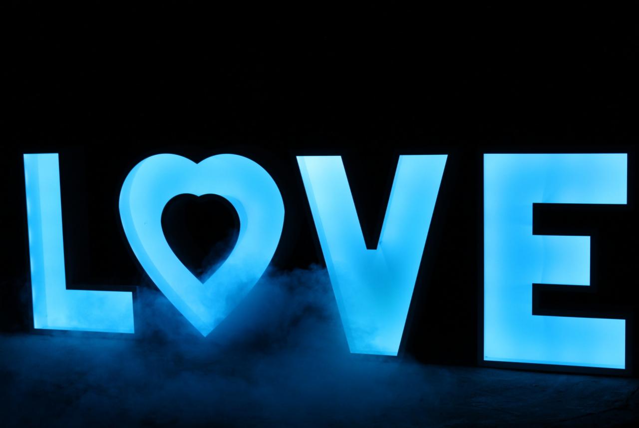 Podświetlany napis LOVE, gdzie litera O zastąpiona jest sercem, otoczony subtelną mgłą, oświetlony na niebiesko na czarnym tle.