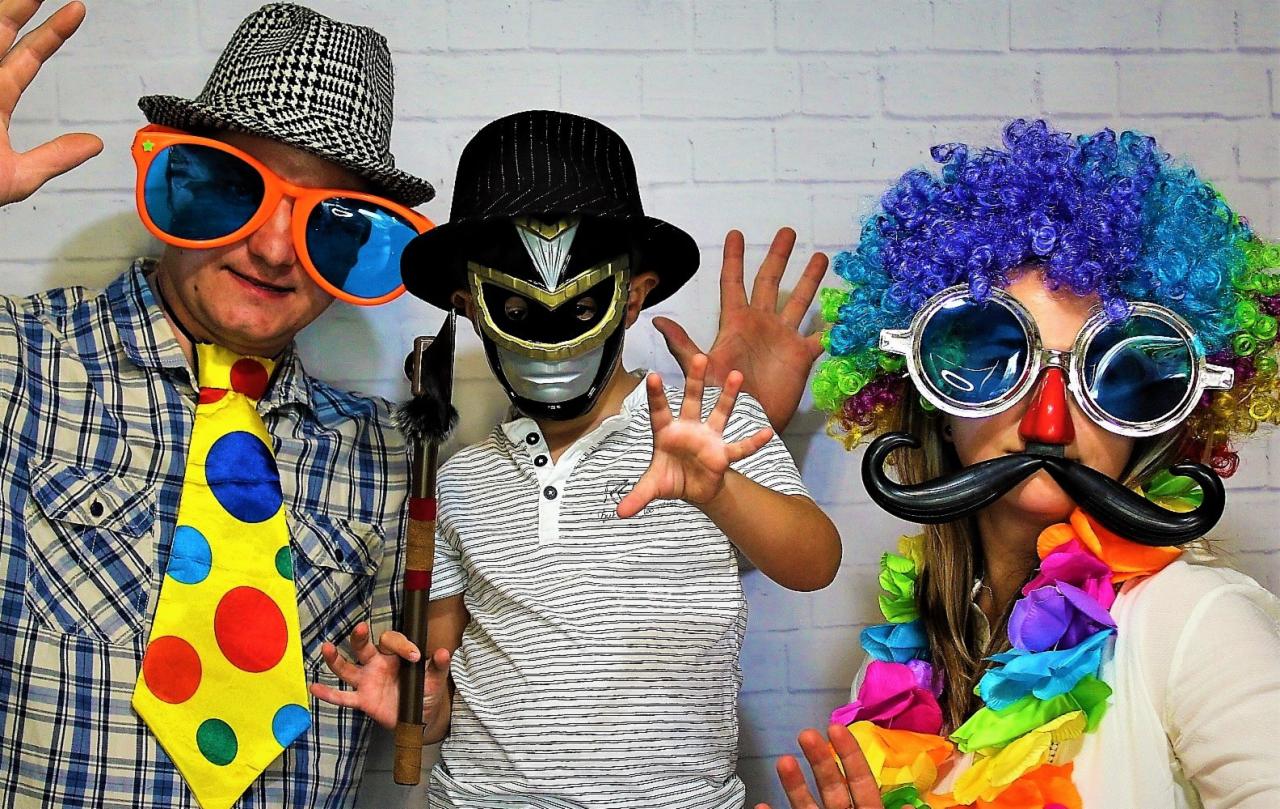 Trzy osoby w przebraniach pozują na tle białej ściany z cegieł: mężczyzna w kraciastej koszuli i dużych okularach, dziecko w masce superbohatera i kobieta w kolorowej peruce i okularach z wąsami.
