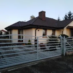 Nowoczesne, poziome przęsła ogrodzeniowe z metalu w kolorze szarym, zabezpieczające posesję z domem jednorodzinnym w tle.