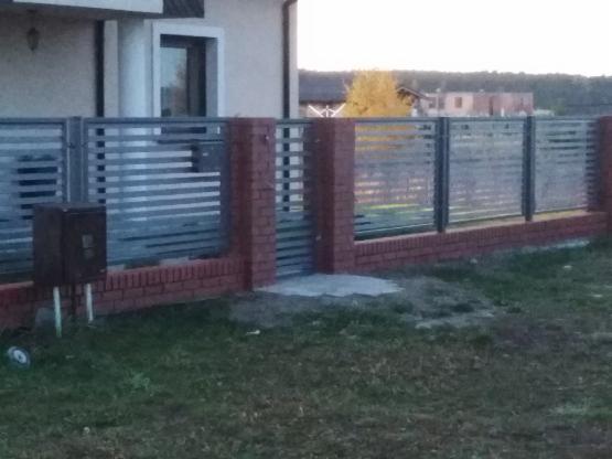 Nowoczesne ogrodzenie z poziomych, metalowych paneli, osadzone na niskim murku z czerwonej cegły, z widoczną skrzynką na listy przy jednym z segmentów.