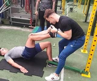 Trener personalny pomaga mężczyźnie w ćwiczeniu unoszenia bioder z jedną nogą na podwyższeniu, używając gumy oporowej, w tle widoczny sprzęt do ćwiczeń.