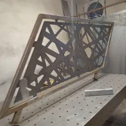 Metalowa konstrukcja ażurowa z wzorem geometrycznym, spawana na stole spawalniczym z widocznymi śladami spawania i niedoskonałościami, ustawiona ukośnie w warsztacie.