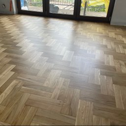 Kacper Cyklinowanie Kacper Jechna - Jasna drewniana podłoga ułożona w klasyczną jodełkę, z widocznymi refleksami światła dziennego, w pomieszczeniu z dużymi drzwiami balkonowymi.