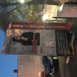 Billboard wyborczy z wizerunkiem Gabrieli Andrzejewskiej, kandydatki na Burmistrza Miasta Kościana, ustawiony przy ulicy, widoczny fragment zaparkowanego samochodu i drzewo w tle.