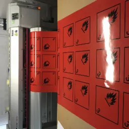 Proces drukowania czerwonych naklejek ostrzegawczych z symbolem łatwopalności na maszynie drukarskiej.