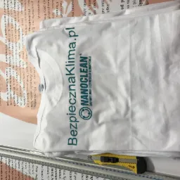 Biała koszulka z nadrukiem 'Bezpiecznaklima.pl' i logo Nanoclean leżąca na tle zadrukowanej kartki papieru, obok żółty nóż introligatorski.