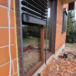 Montaż żaluzji fasadowych zewnętrznych, widoczne przewody i gruz budowlany, okno z brązową ramą, ściana z czerwonej cegły dziurawki.