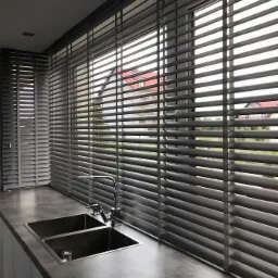 Szare poziome żaluzje drewniane zamontowane w oknach kuchni z widokiem na domy na zewnątrz, widoczne zlewozmywaki ze stali nierdzewnej na blacie.