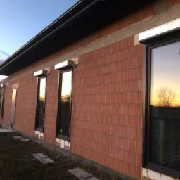 Montaż zewnętrznych rolet okiennych na surowej ścianie budynku z czerwonej cegły, widoczne przewody elektryczne, odbicie zachodu słońca w oknach.