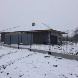 Zimowy krajobraz z nowo postawionym, czarnym ogrodzeniem panelowym na betonowej podmurówce, otaczającym budynek w trakcie budowy, pokryty śniegiem.