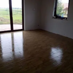 Nowo ułożone, błyszczące panele podłogowe w pustym pokoju z dużym oknem balkonowym i oknem z widokiem na zewnątrz, na parapecie butelka z płynem.