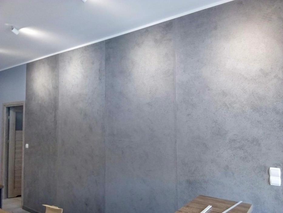 Szara ściana w pokoju z efektem betonu architektonicznego, oświetlona punktowymi lampami sufitowymi, widoczny fragment drzwi i włącznik światła.