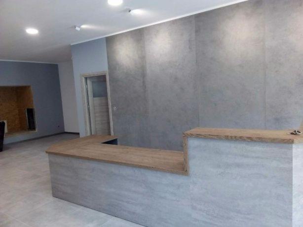 Szara ściana z efektem betonu w wyremontowanym biurze, widoczna drewniana lada recepcyjna i fragmenty innych pomieszczeń w tle.