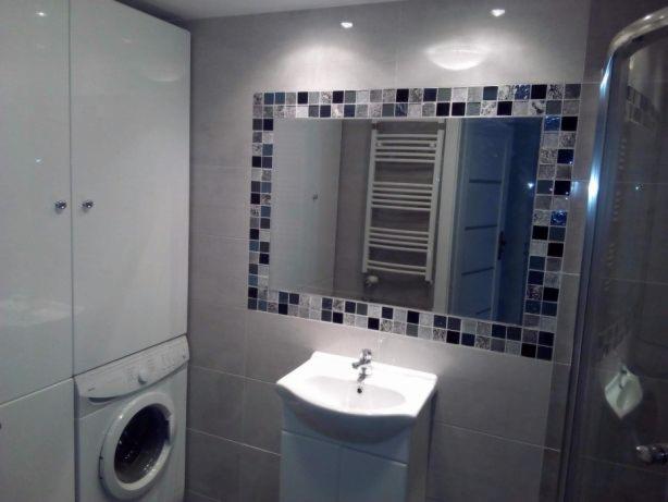 Nowoczesna łazienka w odcieniach szarości z lustrem obramowanym mozaiką, białą umywalką i pralką w zabudowie.