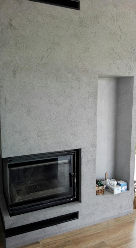 Ściana z kominkiem wykończona szarą, dekoracyjną fakturą, obok wnęka z drobnymi przedmiotami, widoczne panele podłogowe.