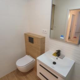 Specmar - Nowoczesna łazienka po remoncie: podwieszana toaleta z drewnianą obudową, biała umywalka z czarną armaturą i szafką z dwoma szufladami, lustro z wyświetlaczem, płytki imitujące drewno na podłodze.