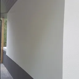 Fragment elewacji domu z widocznym cokołem w kolorze grafitowym oraz białą ścianą o zróżnicowanej fakturze, zarys drzew w tle.