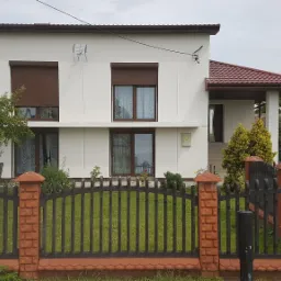 Biały dom jednorodzinny z brązowymi roletami i czerwoną dachówką, widziany zza czarnego ogrodzenia z ceglanymi słupkami.