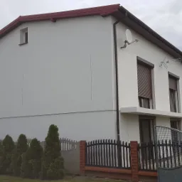 Dwukondygnacyjny dom z ocieploną, białą elewacją, brązowymi roletami w oknach i ciemnoczerwonym dachem, widoczny zza ogrodzenia z tujami.