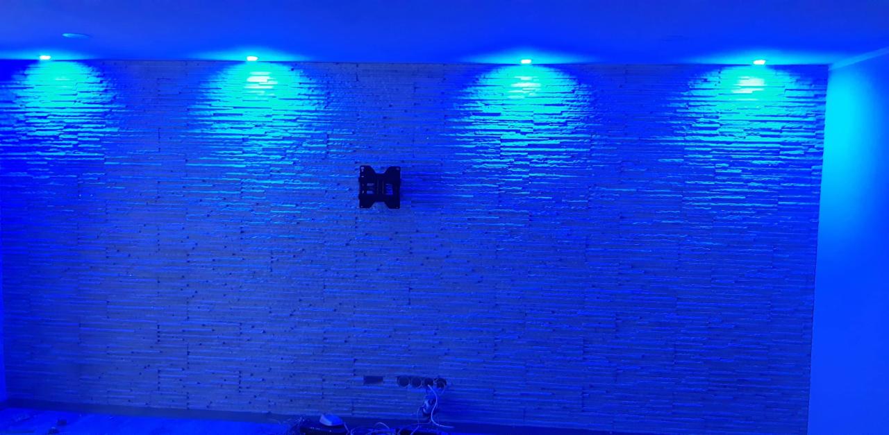 Ściana wykończona dekoracyjną okładziną kamienną, oświetlona niebieskim światłem LED, z zamontowanym uchwytem do telewizora i widocznymi gniazdkami elektrycznymi u dołu.