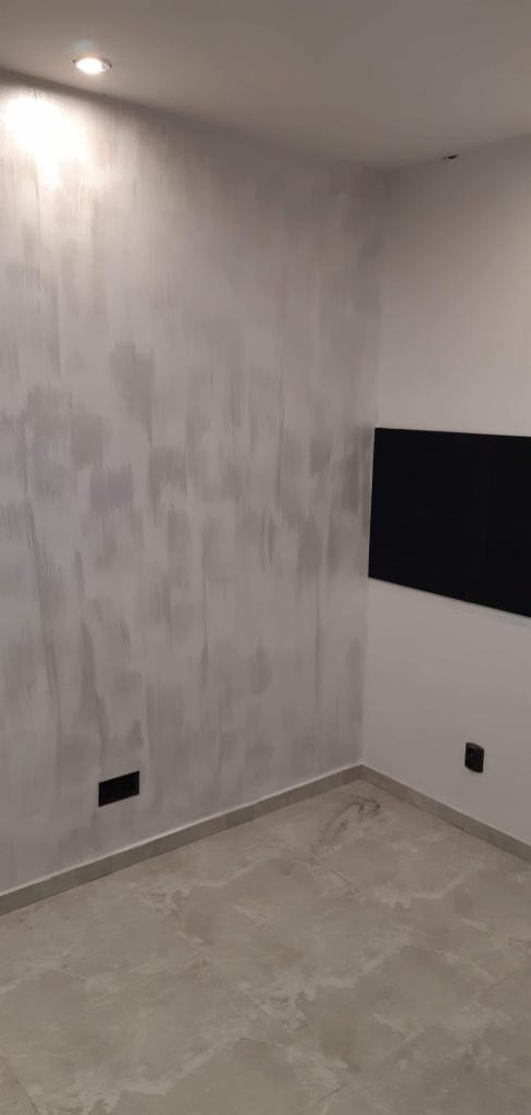 Ściana w pokoju po malowaniu z efektem dekoracyjnym, widoczny czarny panel na ścianie oraz szara podłoga z płytek imitujących marmur.