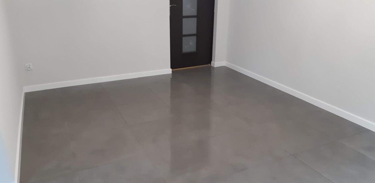 Puste, wyremontowane pomieszczenie z szarymi płytkami podłogowymi, białymi ścianami i ciemnymi drzwiami z przeszkleniami.