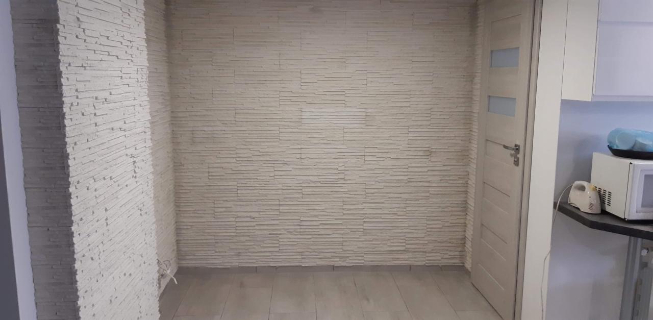 Wykończona ściana w salonie z dekoracyjnym panelem imitującym kamień, jasna podłoga, białe drzwi z szybami, fragment blatu kuchennego z mikrofala i mikserem w tle.