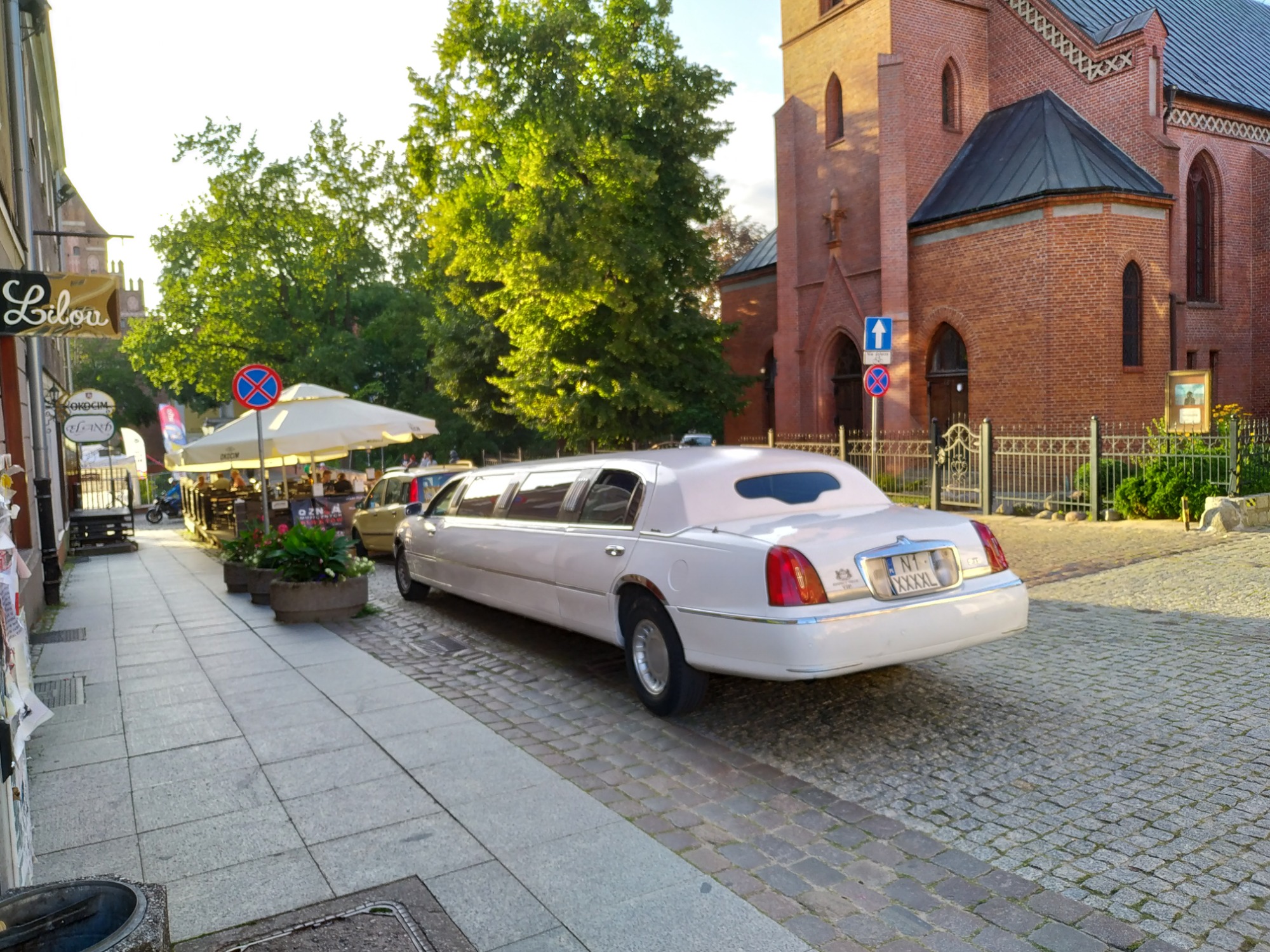 Długa, biała limuzyna zaparkowana na brukowanej ulicy w pobliżu ceglanego kościoła i ogródka restauracyjnego.