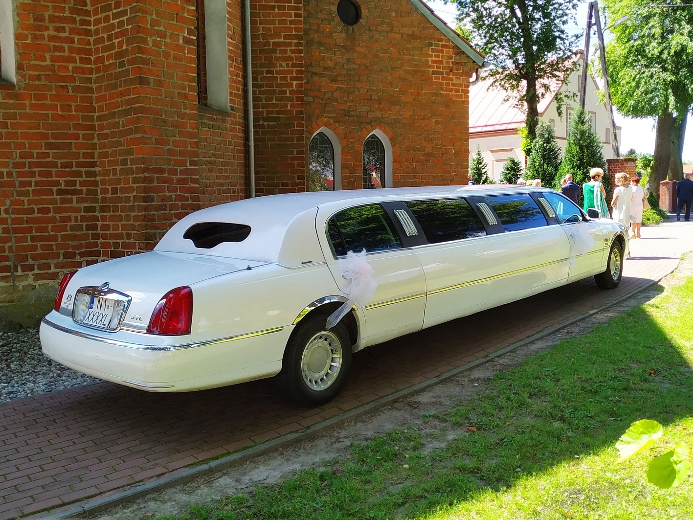 Biała limuzyna Lincoln z dekoracją kwiatową na klamce, zaparkowana na tle ceglanego budynku z gotyckimi oknami i fragmentem zieleni.