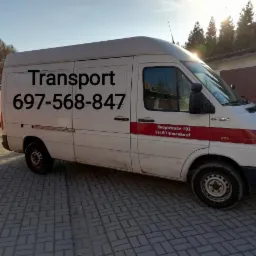 Biały samochód dostawczy z napisem 'Transport' i numerem telefonu zaparkowany na kostce brukowej przed garażami.