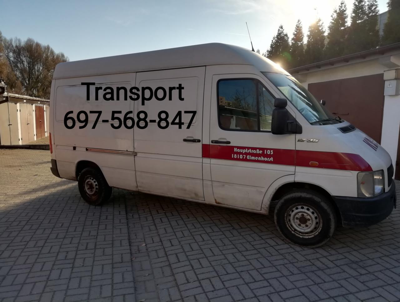 Biały samochód dostawczy z napisem 'Transport' i numerem telefonu zaparkowany na kostce brukowej przed garażami.