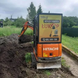 Pomarańczowa minikoparka Hitachi z tablicą reklamową 'Usługi Minikoparką ŁUKAP' w trakcie pracy, nabiera ziemię z wykopu na tle zieleni i zabudowań.