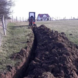 Wąski wykop w ziemi wykonany minikoparką na tle wiejskiego krajobrazu z domem i ogrodzeniem.