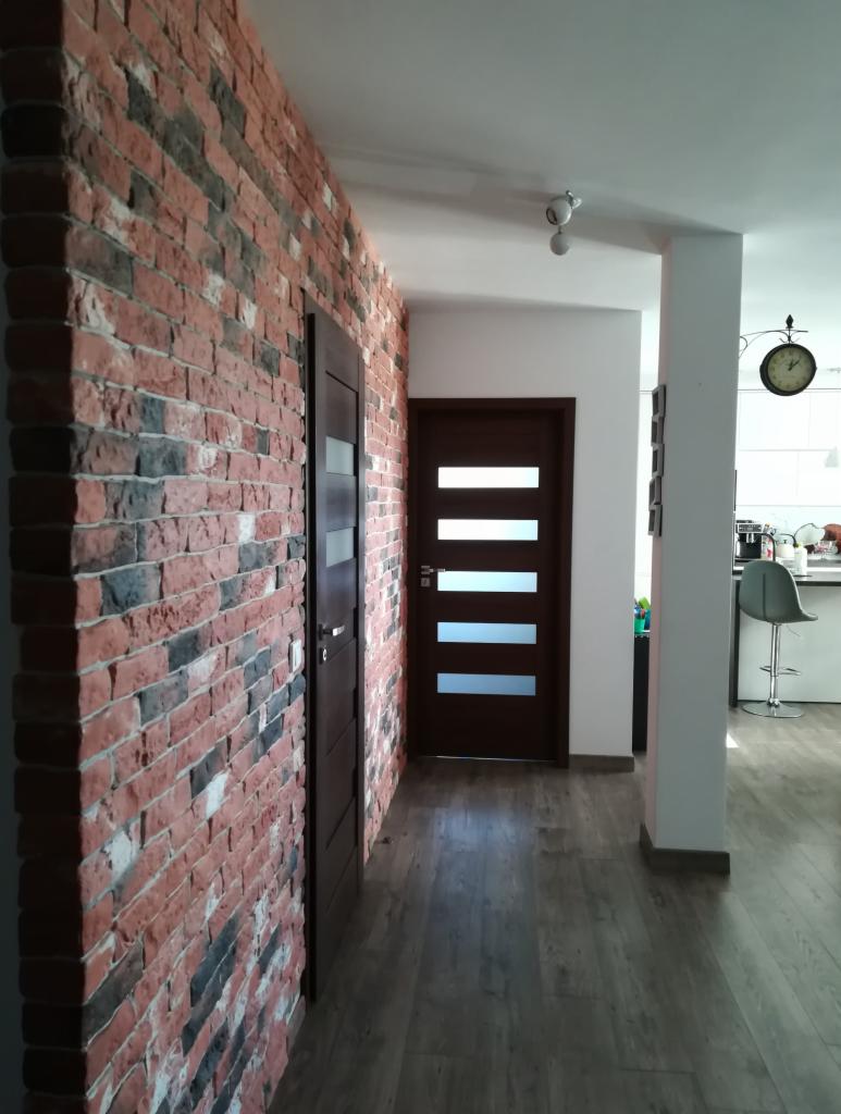 Korytarz z dekoracyjną ścianą z cegły, brązowymi drzwiami z poziomymi przeszkleniami i podłogą imitującą drewno.